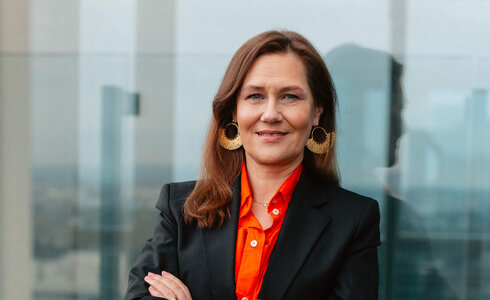 Eine Frau in formeller Kleidung lächelt mit den Armen verschränkt auf einer Terrasse.