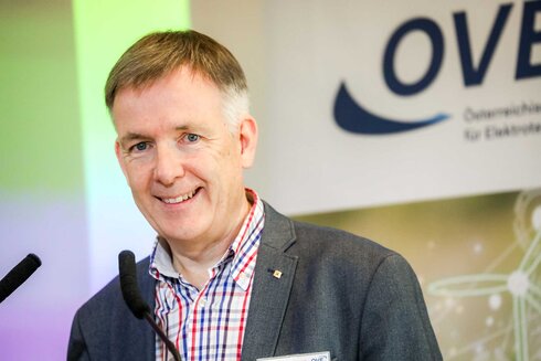 Ein Mann steht lächelnd vor einem Podium mit einem OVE-Logo.