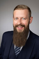 Lächelnder Mann mit Bart, Anzug und Krawatte vor neutralem Hintergrund.