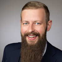 Lächelnder Mann mit Bart, Anzug und Krawatte vor neutralem Hintergrund.