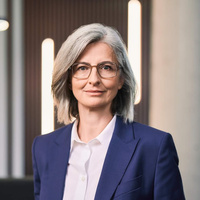 Eine Frau mit grauen Haaren und Brille trägt einen blauen Blazer.