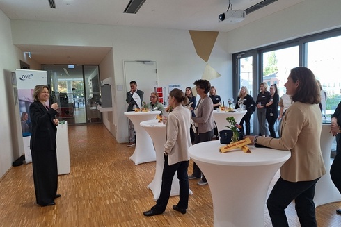 Eine Gruppe von Menschen steht in einem modernen Raum und hört einer Präsentation zu.