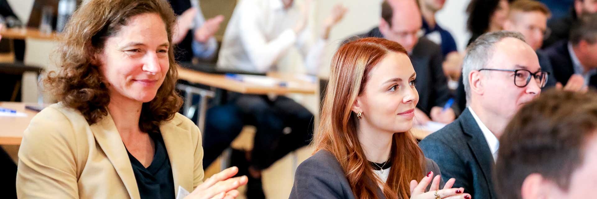 Zwei Frauen im Vordergrund applaudieren in einer lebhaften Veranstaltung mit Publikum.