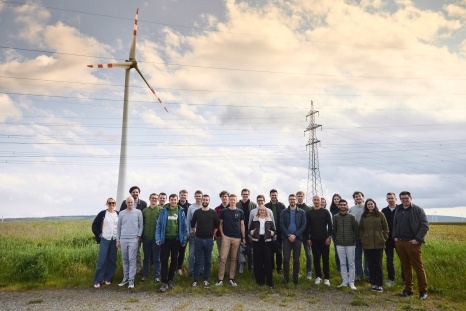 Gruppe von Menschen steht vor einem Windrad und Strommasten in einer Landschaft.