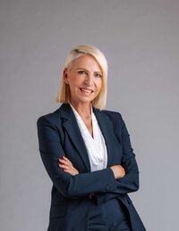 Professionelle Frau in Anzug, lächelnd, mit verschränkten Armen vor neutralem Hintergrund.
