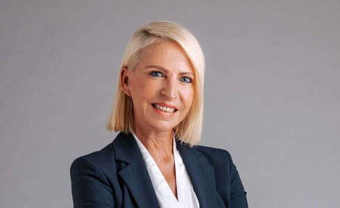 Professionelle Frau in Anzug, lächelnd, mit verschränkten Armen vor neutralem Hintergrund.