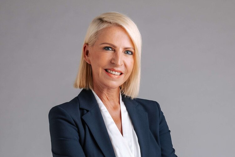 Professionelle Frau in Anzug, lächelnd, mit verschränkten Armen vor neutralem Hintergrund.