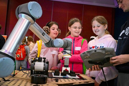 Mädchen beobachten einen Roboterarm und diskutieren dessen Funktionen auf einer Veranstaltung.