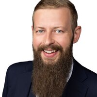 Lächelnder Mann mit Bart in Anzug und Krawatte auf weißem Hintergrund.