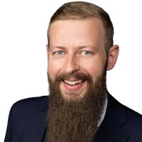Lächelnder Mann mit Bart in Anzug und Krawatte auf weißem Hintergrund.