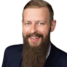 Lächelnder Mann mit Bart in Anzug und Krawatte auf weißem Hintergrund.