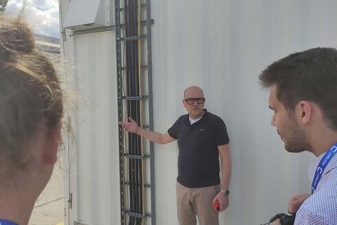 Eine Person erklärt etwas vor einem Container, während Zuhörer aufmerksam zuhören.