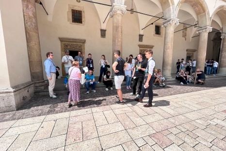 Gruppe von Menschen steht und sitzt in einem Innenhof unter Bögen.
