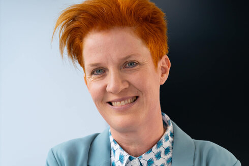 Lächelnde Person mit orangefarbenem Haar und blazer, vor einfarbigem Hintergrund.