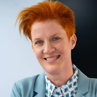 Lächelnde Person mit orangefarbenem Haar und blazer, vor einfarbigem Hintergrund.