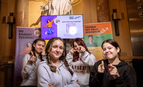 Vier junge Frauen posieren vor einem Bildschirm mit dem Text: „Deine Meinung ist gefragt!“