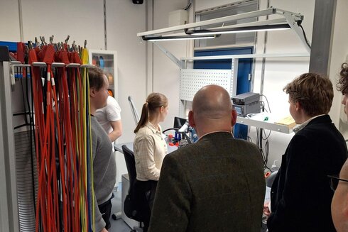 Gruppe von Personen beobachtet eine Präsentation an einem Arbeitsplatz mit technischem Equipment.