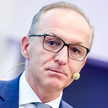 Ein Mann mit Brille und Mikrofon am Anzug, schaut ernst in die Kamera.
