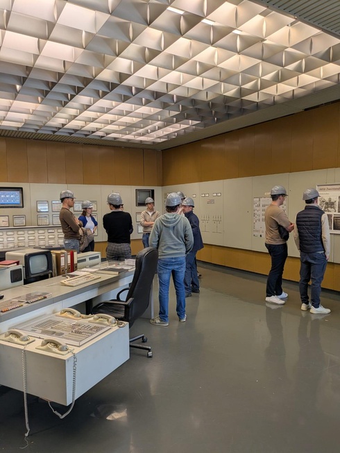 Eine Gruppe von Menschen in einem historischen Kontrollraum, Geräte und Informationsanzeigen sind sichtbar.