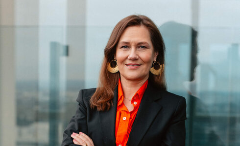 Eine Frau in formeller Kleidung lächelt mit den Armen verschränkt auf einer Terrasse.