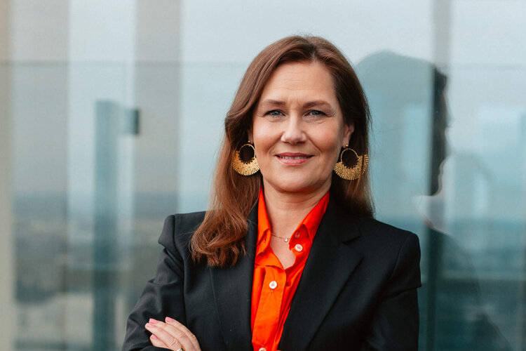 Eine Frau in formeller Kleidung lächelt mit den Armen verschränkt auf einer Terrasse.