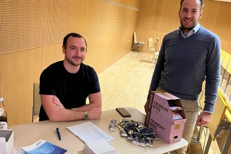 Zwei Männer stehen an einem Tisch mit Snacks und Informationsmaterial.