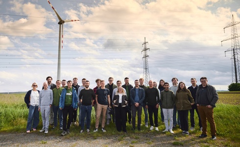 Gruppe von Menschen steht im Freien neben einem Windrad und Strommasten.
