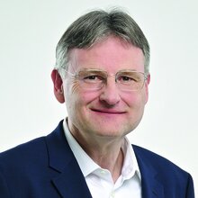 Porträt eines Mannes mit Brille, lächelnd, im Anzug vor neutralem Hintergrund.