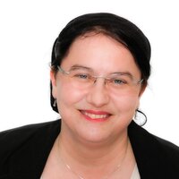 Eine lächelnde Frau mit Brille und schwarzem Blazer auf hellem Hintergrund.