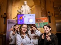 Vier junge Frauen posieren vor einem Bildschirm mit dem Motto „Deine Meinung ist gefragt“.