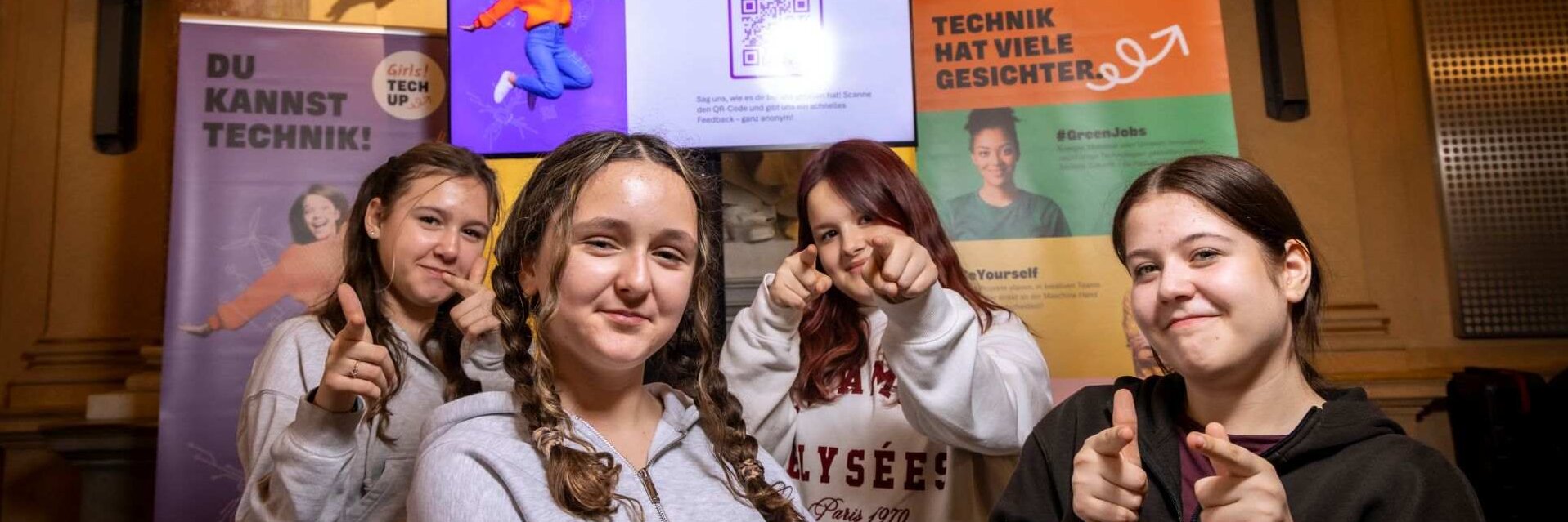 Vier junge Frauen posieren vor einem Bildschirm mit dem Motto „Deine Meinung ist gefragt“.