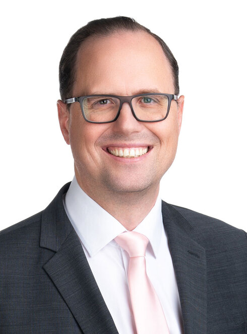 Ein lächelnder Mann in Anzug mit Brille und hellrosa Krawatte.