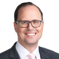 Ein lächelnder Mann in Anzug mit Brille und hellrosa Krawatte.