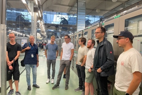 Eine Gruppe von acht Personen hört einem Mann in einer Fabrikhalle zu.