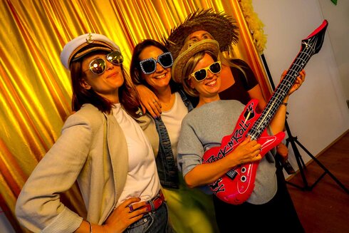 Vier Frauen posieren mit lustigen Accessoires und einer Spielzeuggitarre vor goldenem Hintergrund.