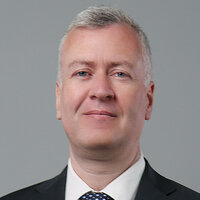 Ein Mann in einem Anzug mit Krawatte lächelt vor einem neutralen Hintergrund.