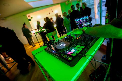 DJ-Setup mit Mischpult, umgeben von Gästen auf einer Veranstaltung.