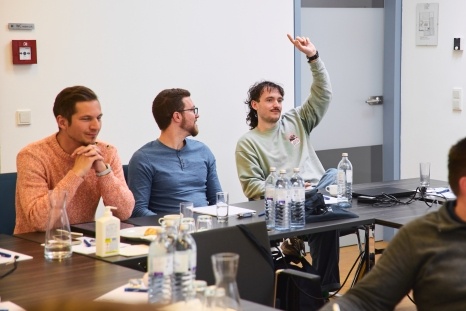 Drei Personen sitzen an einem Tisch, einer hebt die Hand.