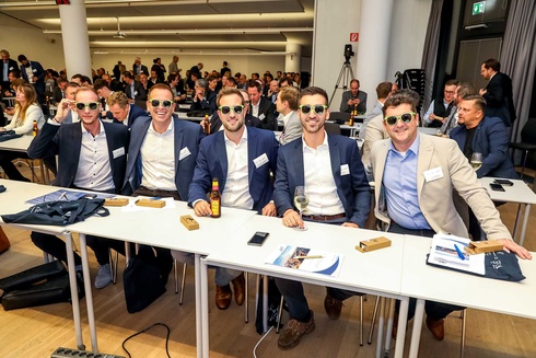 Fünf Männer mit Sonnenbrillen lächeln am Tisch bei einer Veranstaltung.