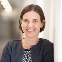 Lächelnde Frau steht an einer Wand in einem hellen Innenraum.