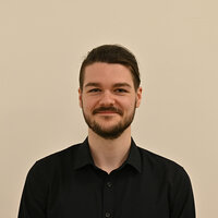 Porträt eines jungen Mannes mit Bart, der lächelt, vor einem neutralen Hintergrund.