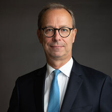 Ein Mann in Anzug und Krawatte mit Brille posiert vor einem dunklen Hintergrund.