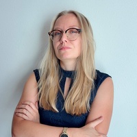 Frau mit langen blonden Haaren und Brille, in dunklem Kleid, steht mit verschränkten Armen.
