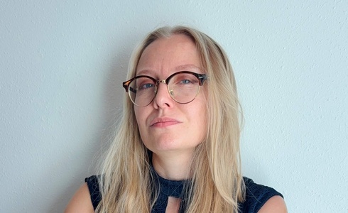 Frau mit langen blonden Haaren und Brille, in dunklem Kleid, steht mit verschränkten Armen.