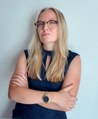 Frau mit langen blonden Haaren und Brille, in dunklem Kleid, steht mit verschränkten Armen.