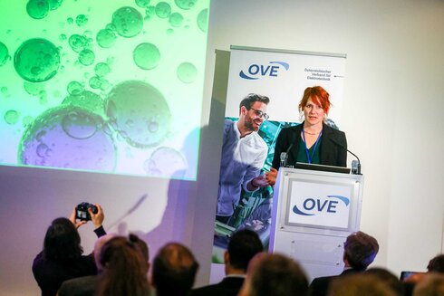 Eine Frau spricht auf einer Konferenz mit einer Grafik im Hintergrund.