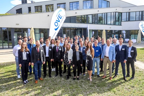 Gruppe von Menschen posiert vor einem Gebäude mit Schildern und Sonnenschirmen.