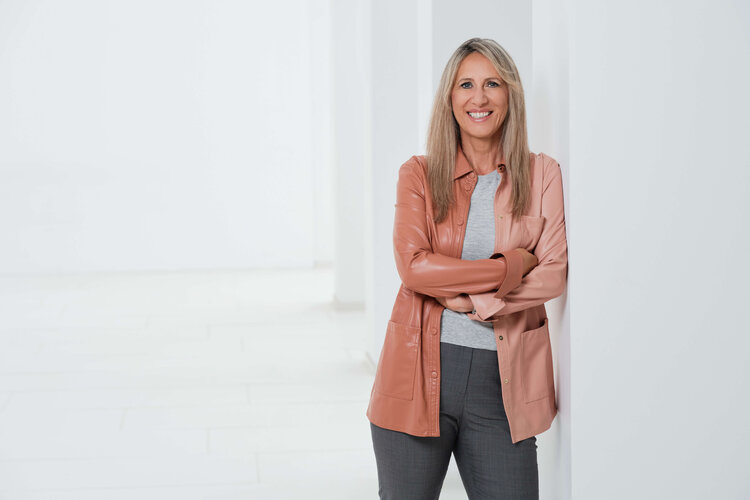 Frau mit blonden Haaren lächelt und steht an einer Wand.