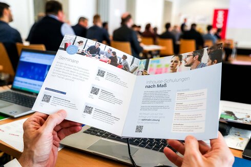 Person hält einen Faltprospekt über Schulungen und Seminare in einem Konferenzraum.