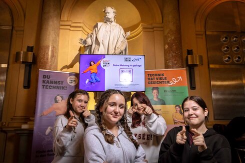 Vier junge Frauen posieren vor einem Bildschirm mit Informationsmaterial zu Technik.